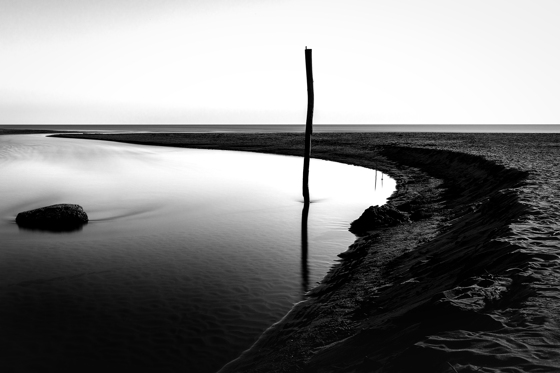 Palo solitario riflesso nell’acqua su una spiaggia deserta, fotografia in bianco e nero minimalista.
