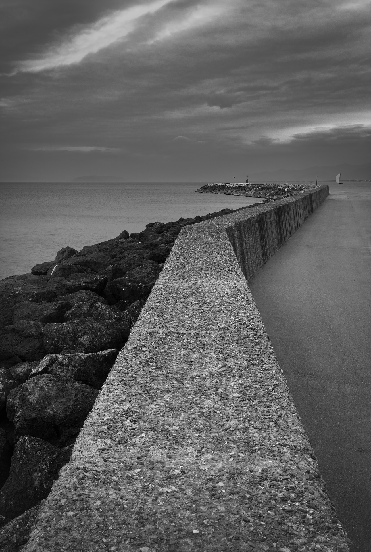 Fotografia minimalista in bianco e nero: un lungo molo di cemento si estende verso il mare sotto un cielo drammatico, enfatizzando isolamento e geometrie urbane.