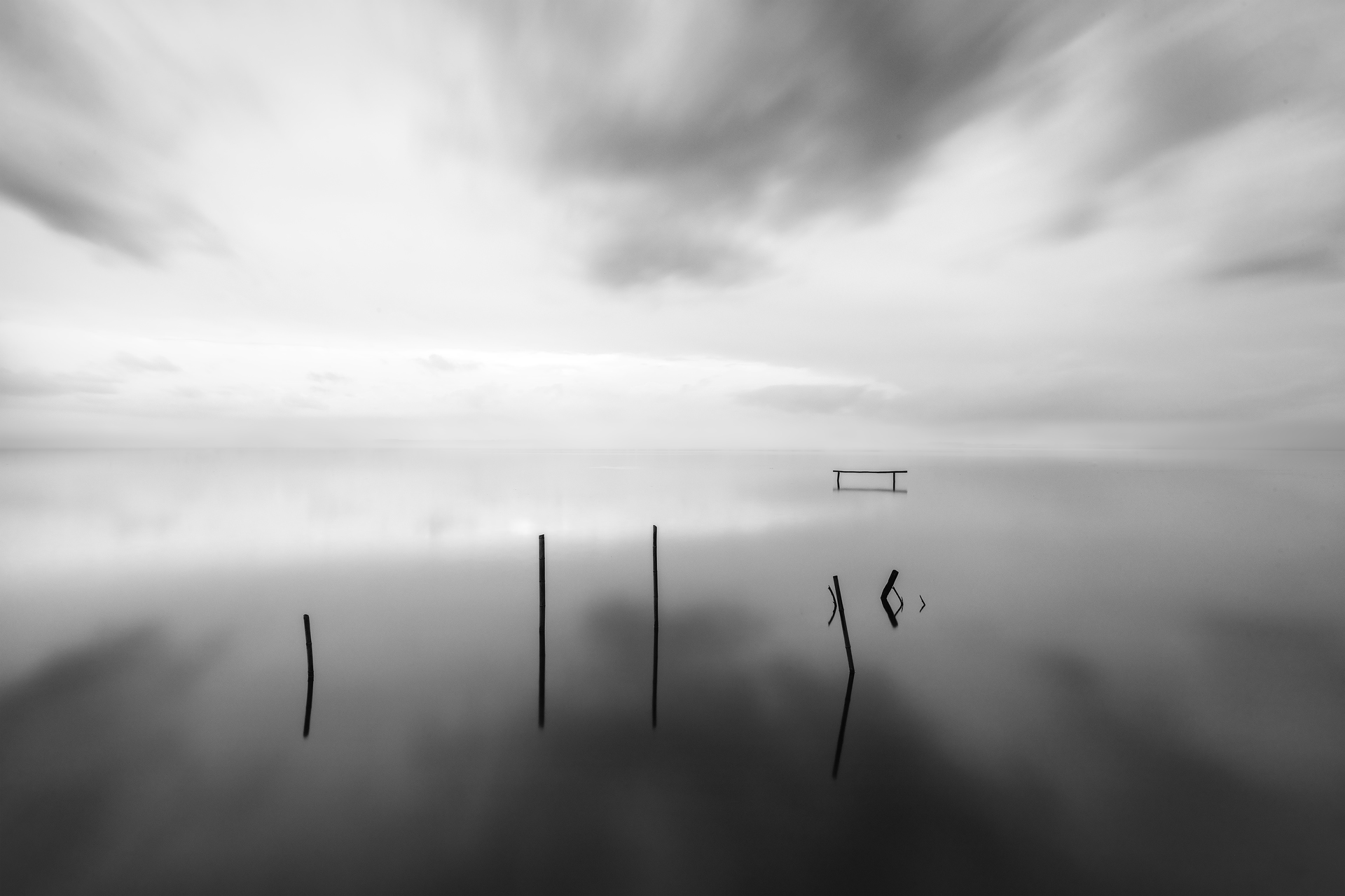 Fotografia minimalista in bianco e nero: un paesaggio acquatico surreale con pali emergenti dall’acqua e nuvole in movimento, evocando silenzio e contemplazione.