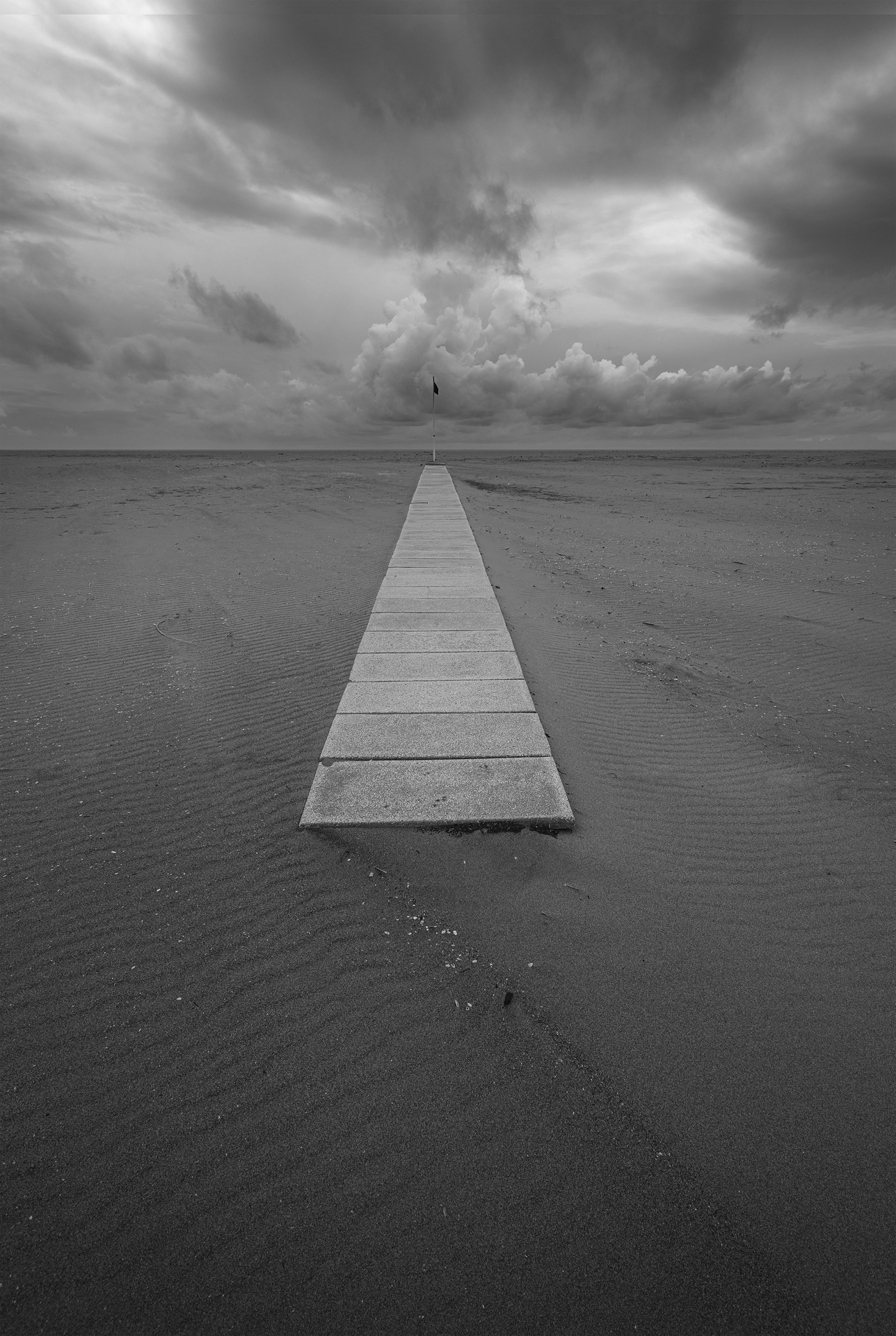 Fotografia minimalista in bianco e nero: un camminamento in cemento conduce l’occhio verso l’orizzonte, attraversando una distesa di sabbia deserta sotto un cielo tempestoso.