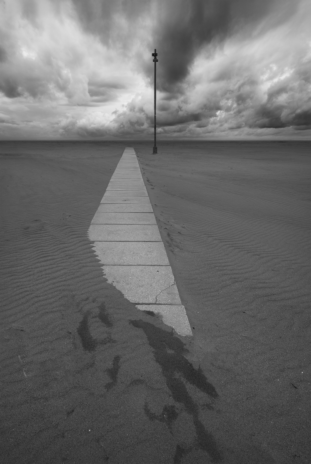 Fotografia minimalista in bianco e nero: una passerella di cemento sepolta dalla sabbia conduce a un megafono solitario sotto un cielo tempestoso.