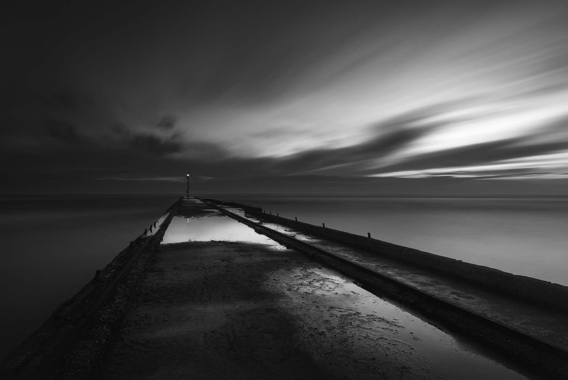 Molo al tramonto in bianco e nero, con cielo mosso e riflessi sull’acqua, fotografia minimalista e drammatica.