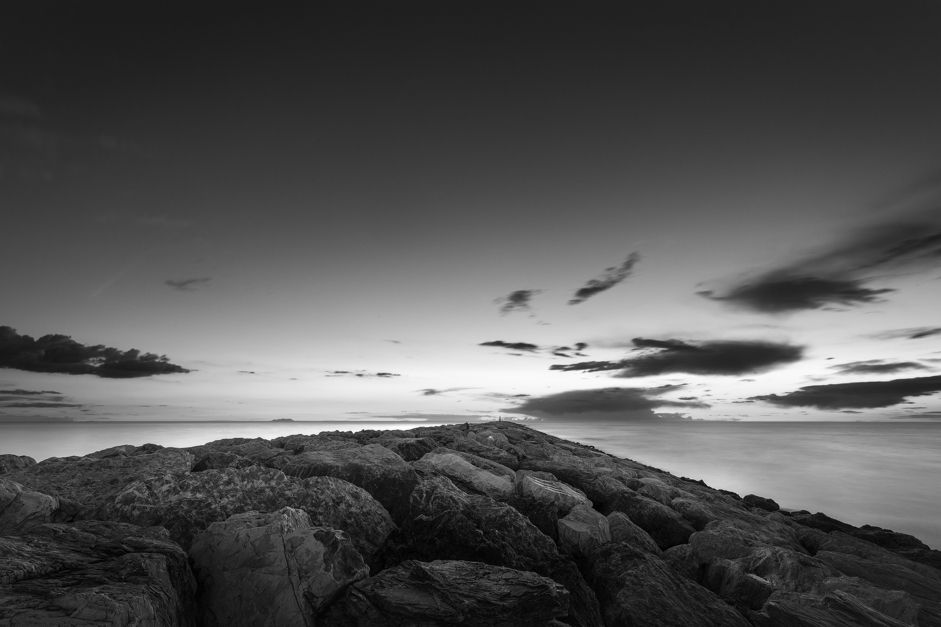 Scogliera che si perde nel mare al tramonto con cielo nuvoloso in bianco e nero.