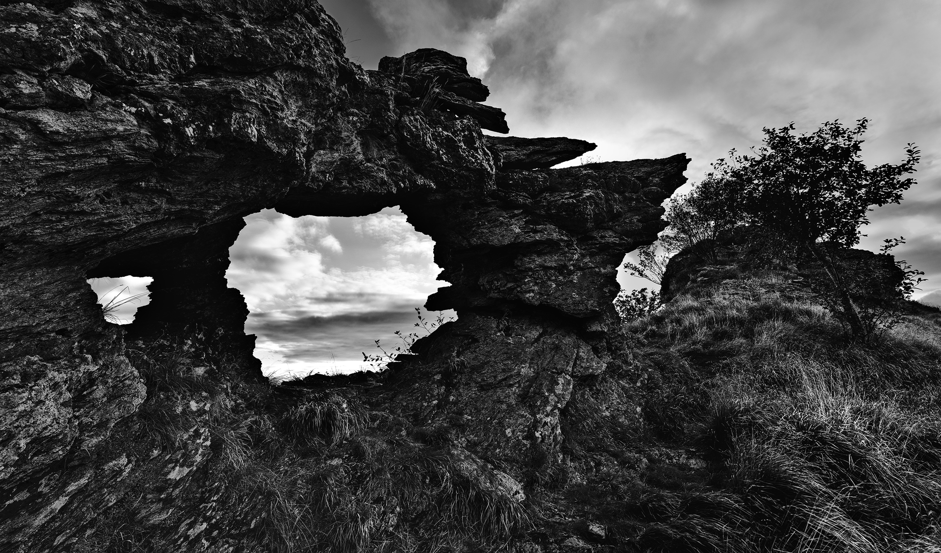 Arco roccioso naturale a Passo Croce in bianco e nero; finestra nella roccia che incornicia cielo e nuvole, texture di pietra e erba mosso-leggera.