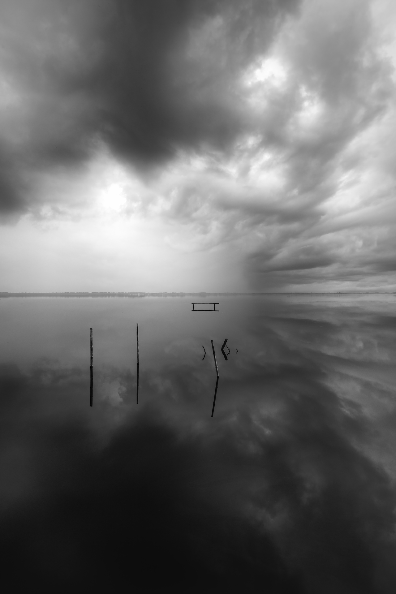 Cielo carico di nuvole sopra un lago immobile. Alcuni pali spezzati emergono dall’acqua, dando un senso di mistero e solitudine.
