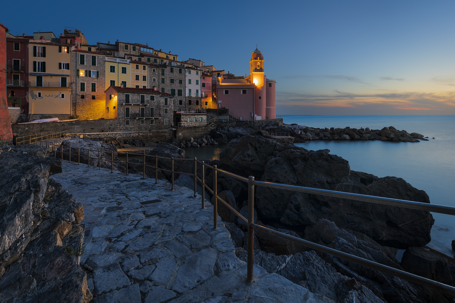 Tramonto dorato su Tellaro. Il borgo illuminato si riflette sul mare calmo, mentre il cielo si sfuma nei toni del blu e dell’arancio.