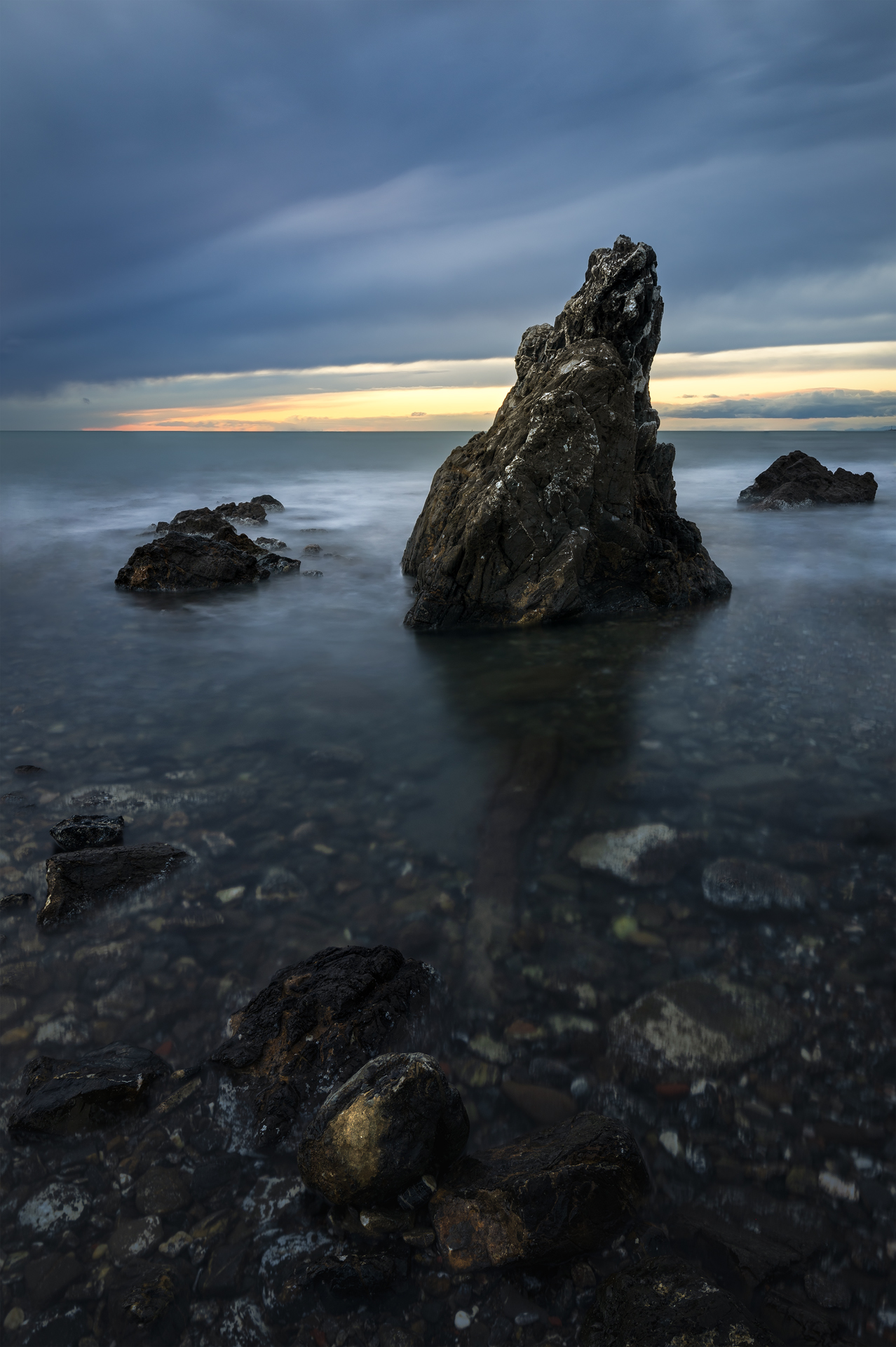 Scoglio solitario in mezzo al mare con il cielo tempestoso. Le onde sfiorano le rocce e la luce dorata dell’alba si riflette sulla superficie.