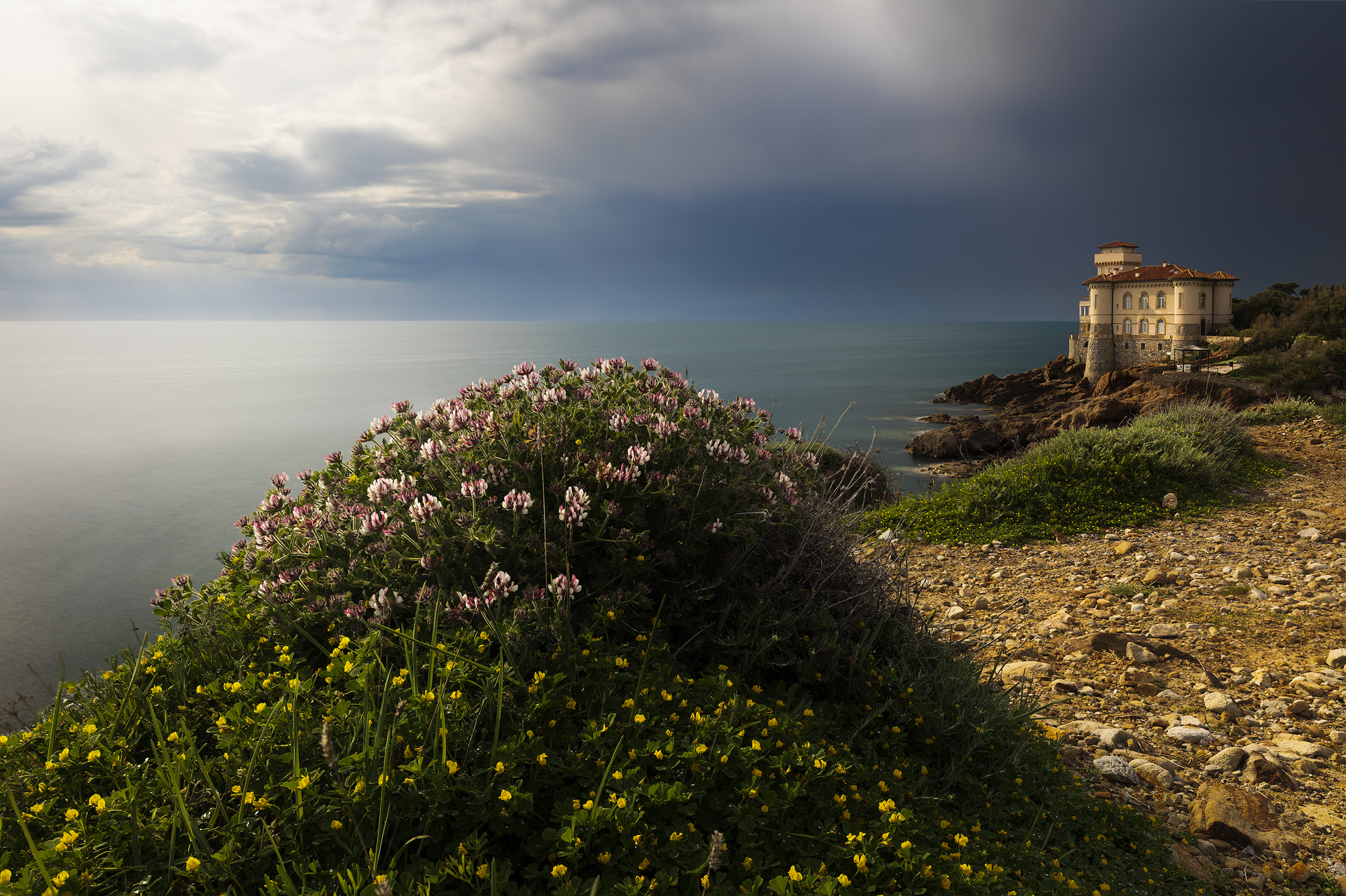 Fotografia panoramica del Castello del Boccale a Calafuria, Livorno, ripreso al tramonto con fiori in primo piano, cielo nuvoloso e mare con effetto seta, realizzata con tecnica di focus stacking ed exposure blending.