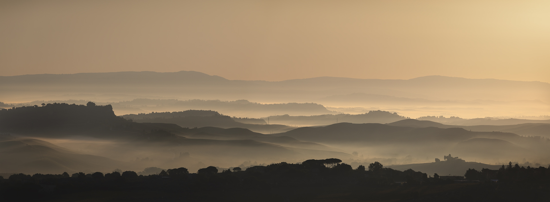 Paesaggio panoramico collinare all'alba con luce dorata e foschia, fotografato da Rivalto, Chianni (PI), Italia.