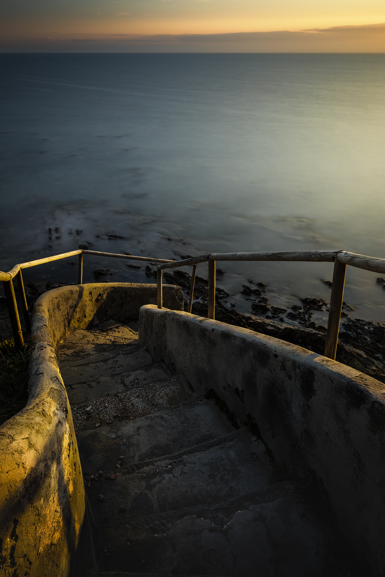 Scala di pietra che scende verso il mare, illuminata dall’ultima luce del giorno. Il tramonto tinge l’orizzonte di tonalità calde e dorate.