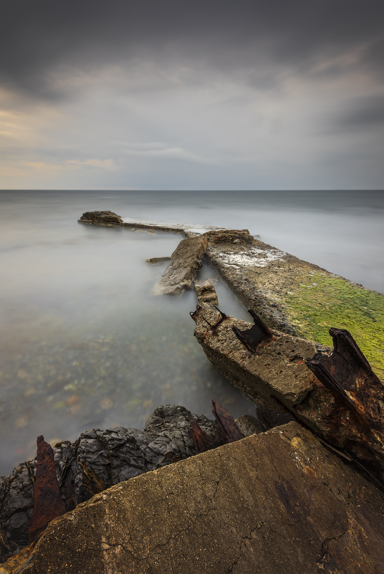 Pontile in rovina che si allunga nel mare calmo. I resti delle travi sommerse creano una geometria suggestiva, con il cielo nuvoloso sullo sfondo.