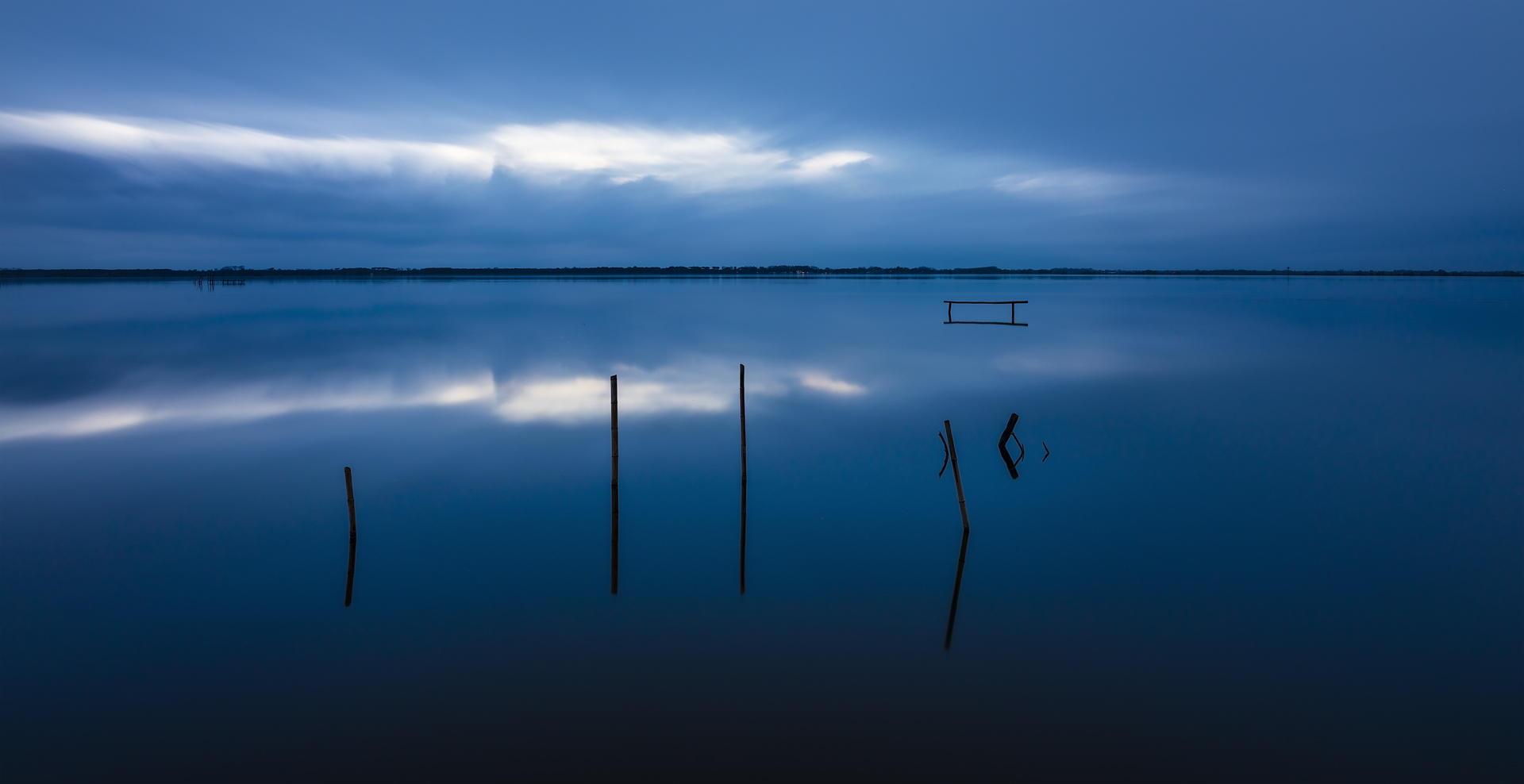Specchio d’acqua blu al Lago di Massaciuccoli al crepuscolo, con pali affioranti e riflessi nelle acque calme. Massaciuccoli, Lucca, Italia