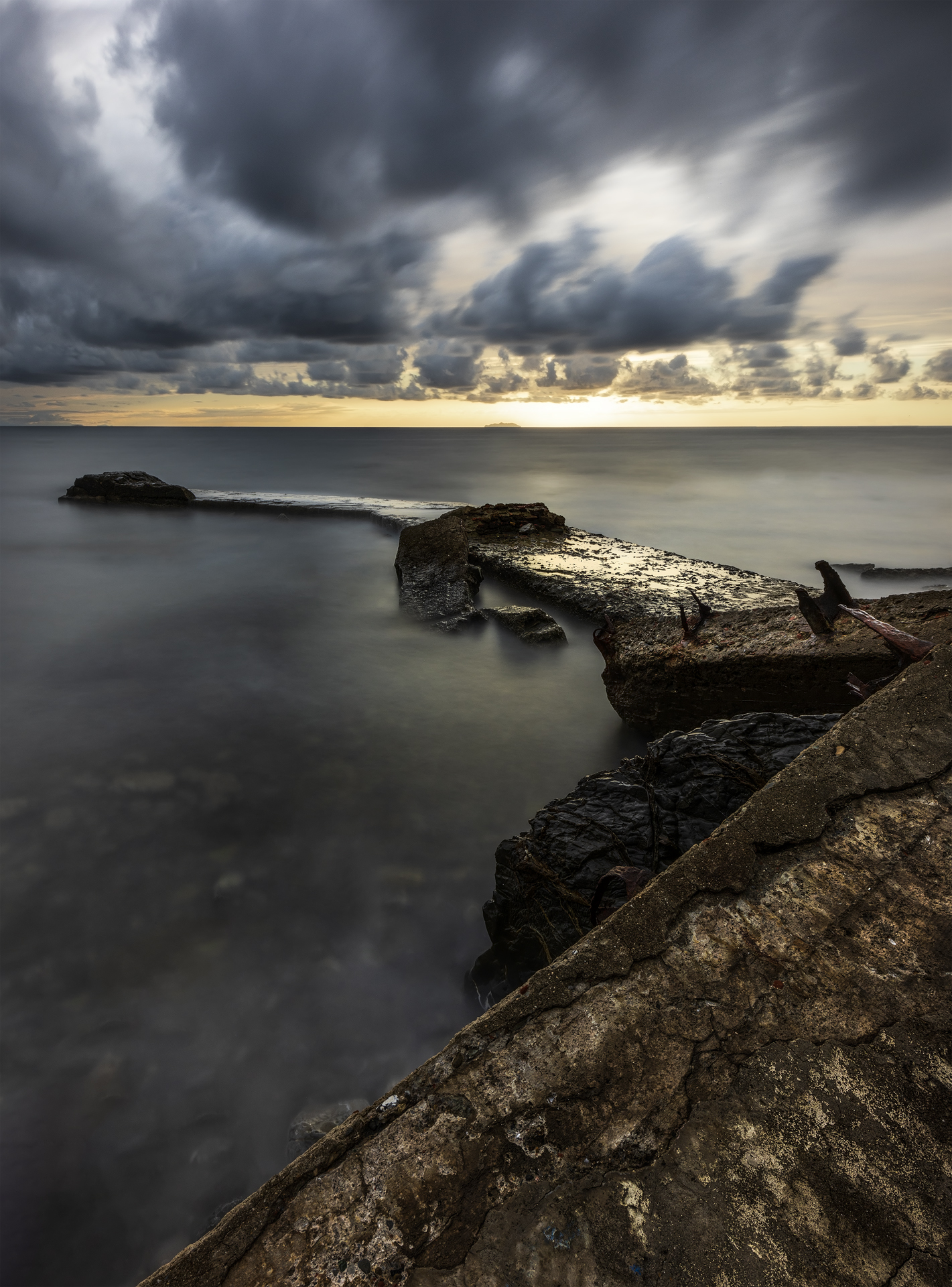 Lunga esposizione al tramonto alla Spiaggetta delle Ville (Livorno): pontile di cemento spezzato che punta l’orizzonte, acqua setosa, cielo nuvoloso drammatico, diagonali in primo piano.