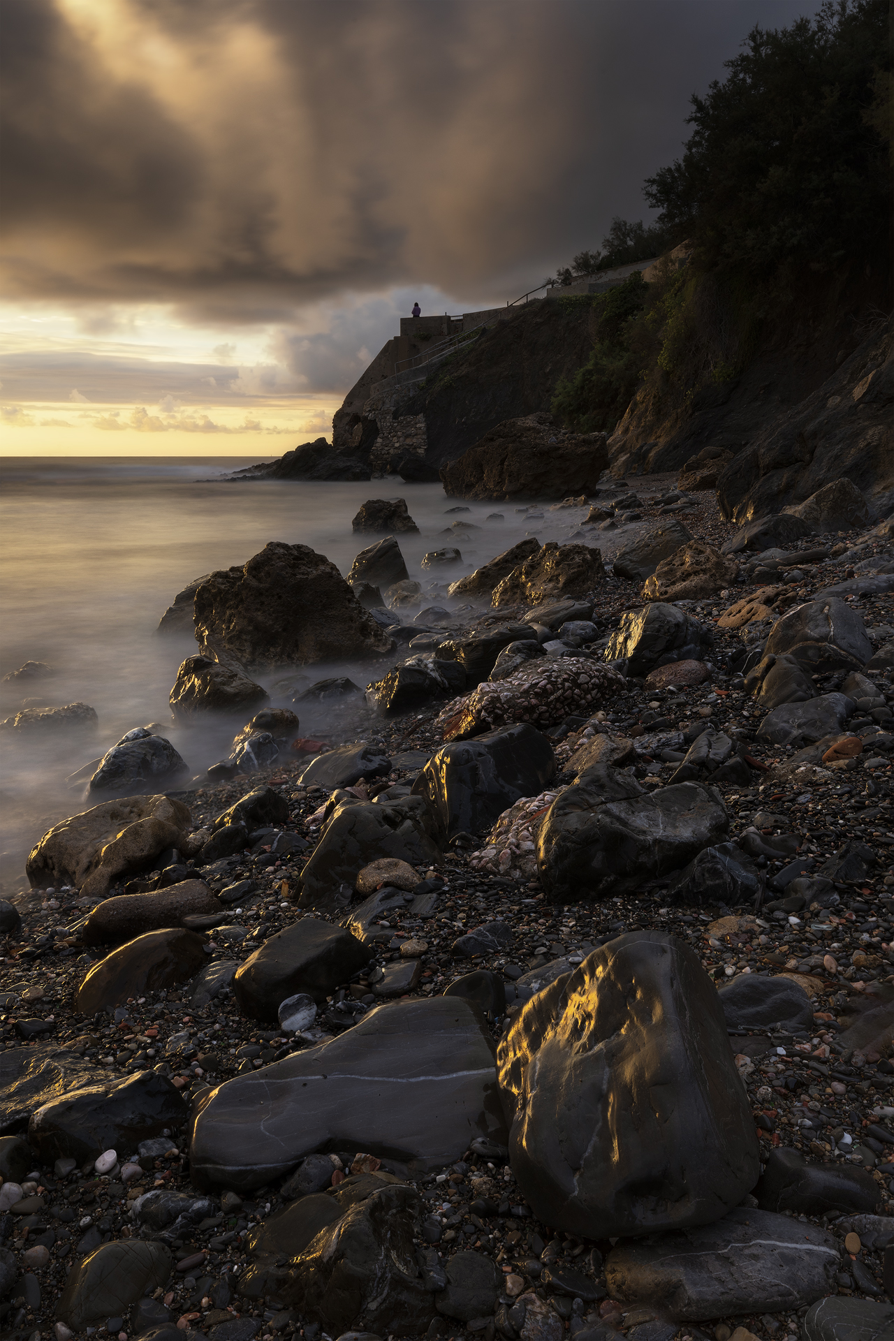 Fotografia di paesaggio al tramonto alla Spiaggetta delle Ville a Livorno: rocce illuminate dalla luce dorata in primo piano, acqua vellutata da lunga esposizione, scogliera con figura solitaria e cielo drammatico con nuvole e raggi di luce.