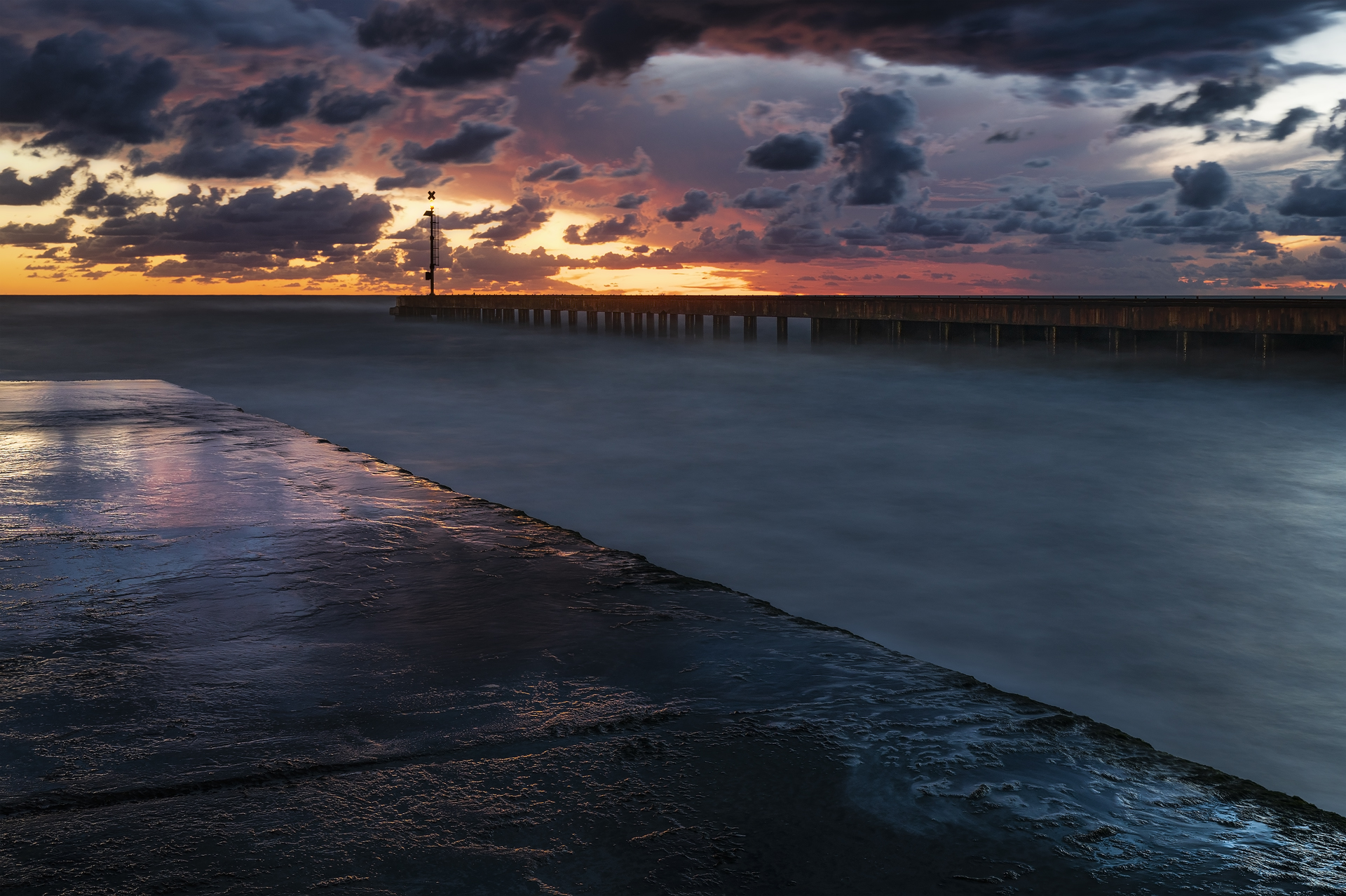 Tramonto al Pontile del Cinquale: molo lucido in primo piano, acqua setosa da lunga esposizione, pontile con faro e cielo drammatico con nuvole e luce calda all’orizzonte.