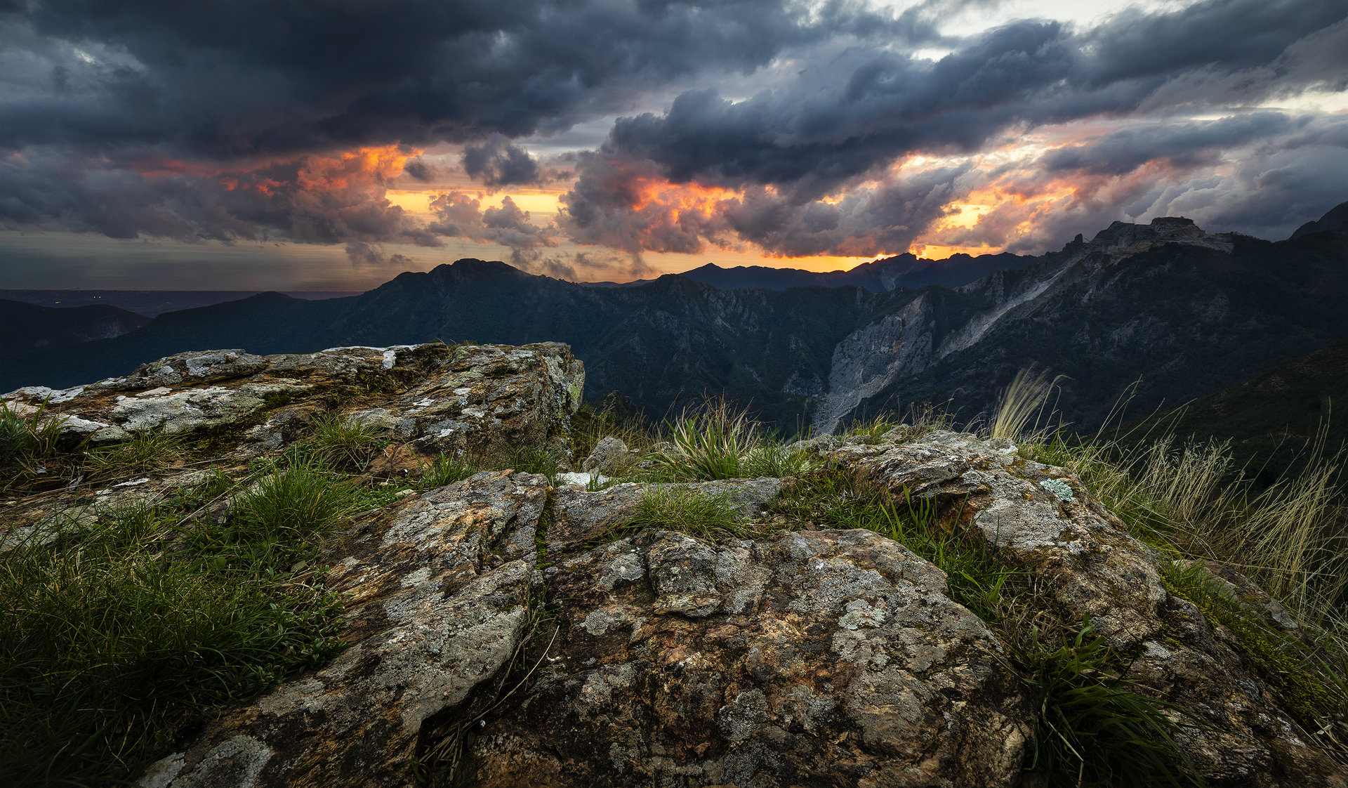 Tramonto al Passo Croce (Alpi Apuane, Lucca): rocce in primo piano, montagne scure e cielo drammatico con nuvole e bagliori arancioni, tecnica focus stacking ed exposure blending.