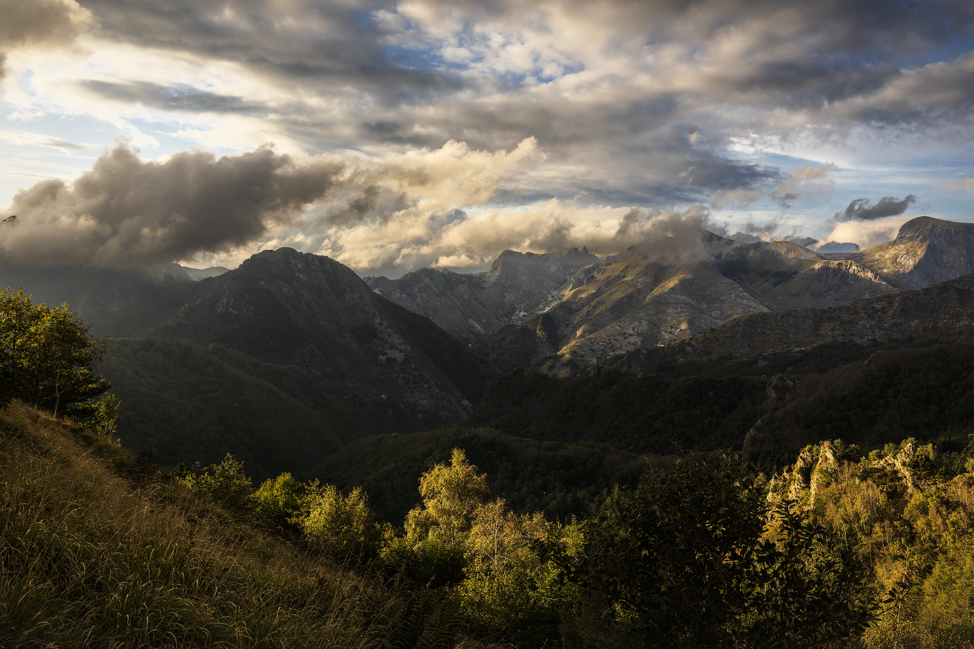 Panorama Apuane al tramonto da Passo Croce: valle in luce radente, creste illuminate e nuvole dinamiche; erba dorata in primo piano, exposure blending naturale.