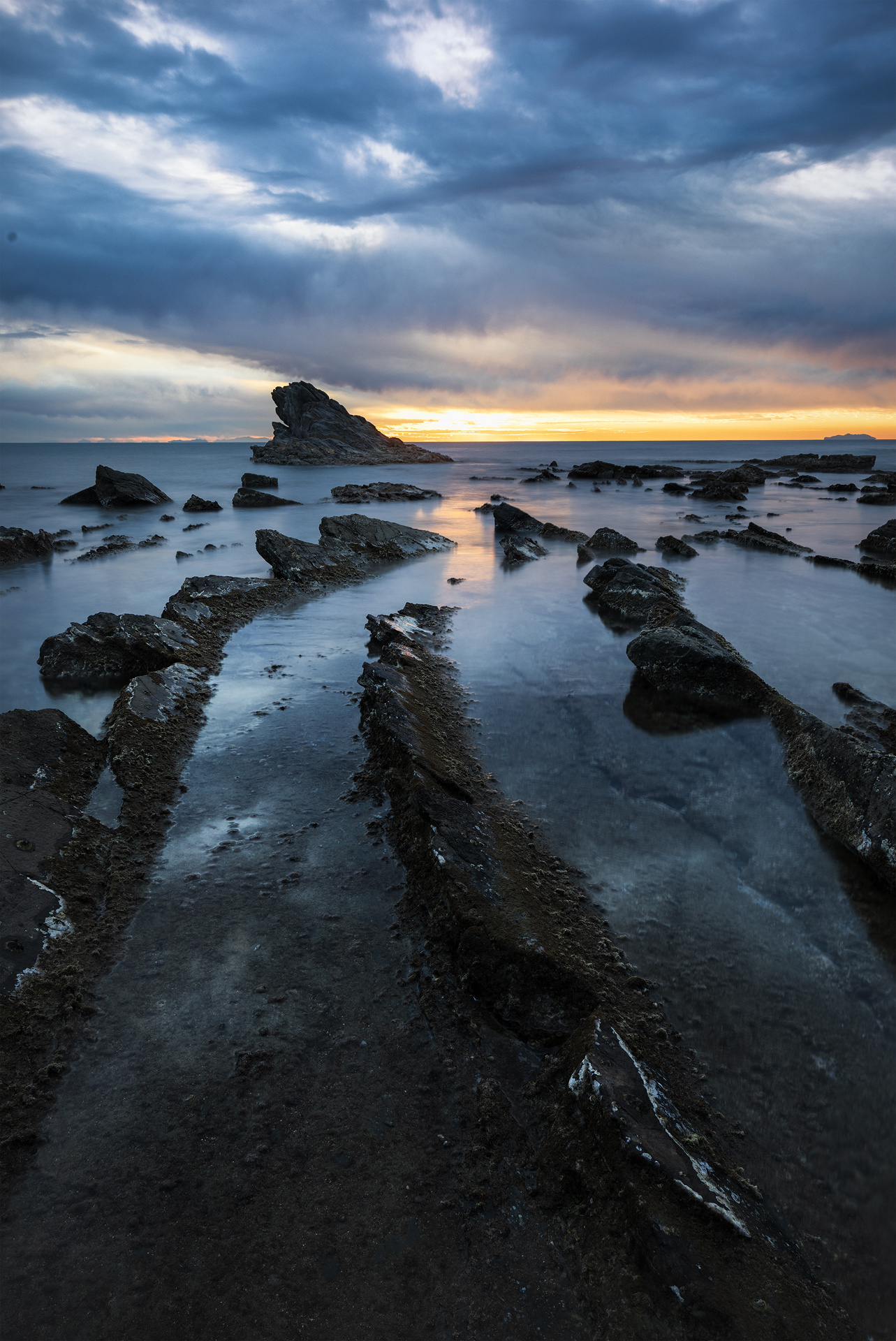 Tramonto a Punta del Casotto, Antignano: rocce a ventaglio in primo piano, acqua setosa da lunga esposizione, isolotto a sinistra e fascia arancio all’orizzonte, focus stacking + exposure blending.