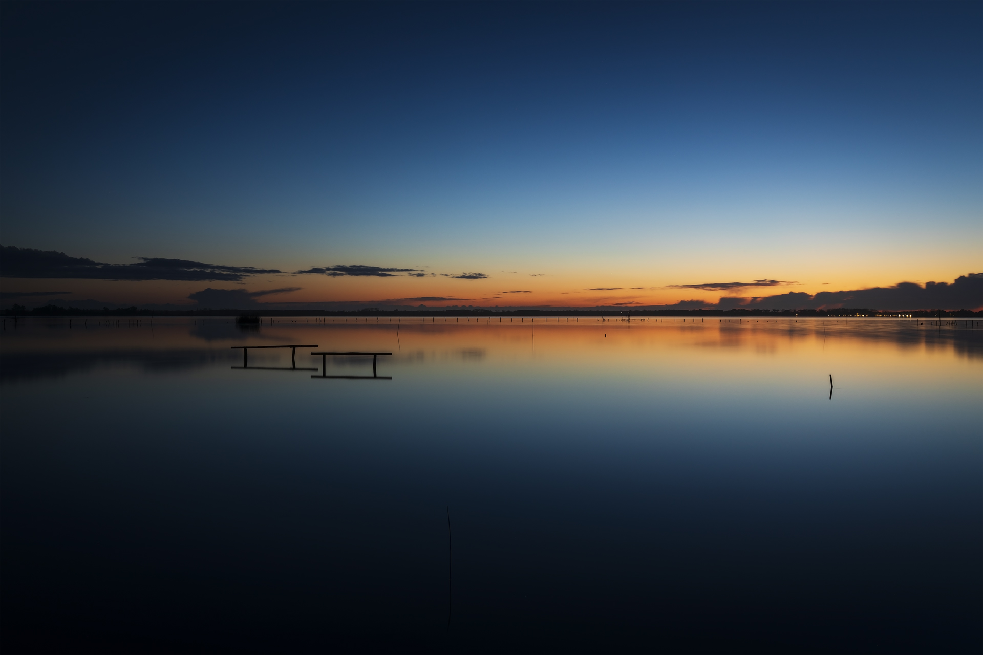 Lunga esposizione su una laguna al tramonto. Il cielo limpido si riflette perfettamente sull’acqua, mentre le sagome di alcuni pali emergono dall’orizzonte.