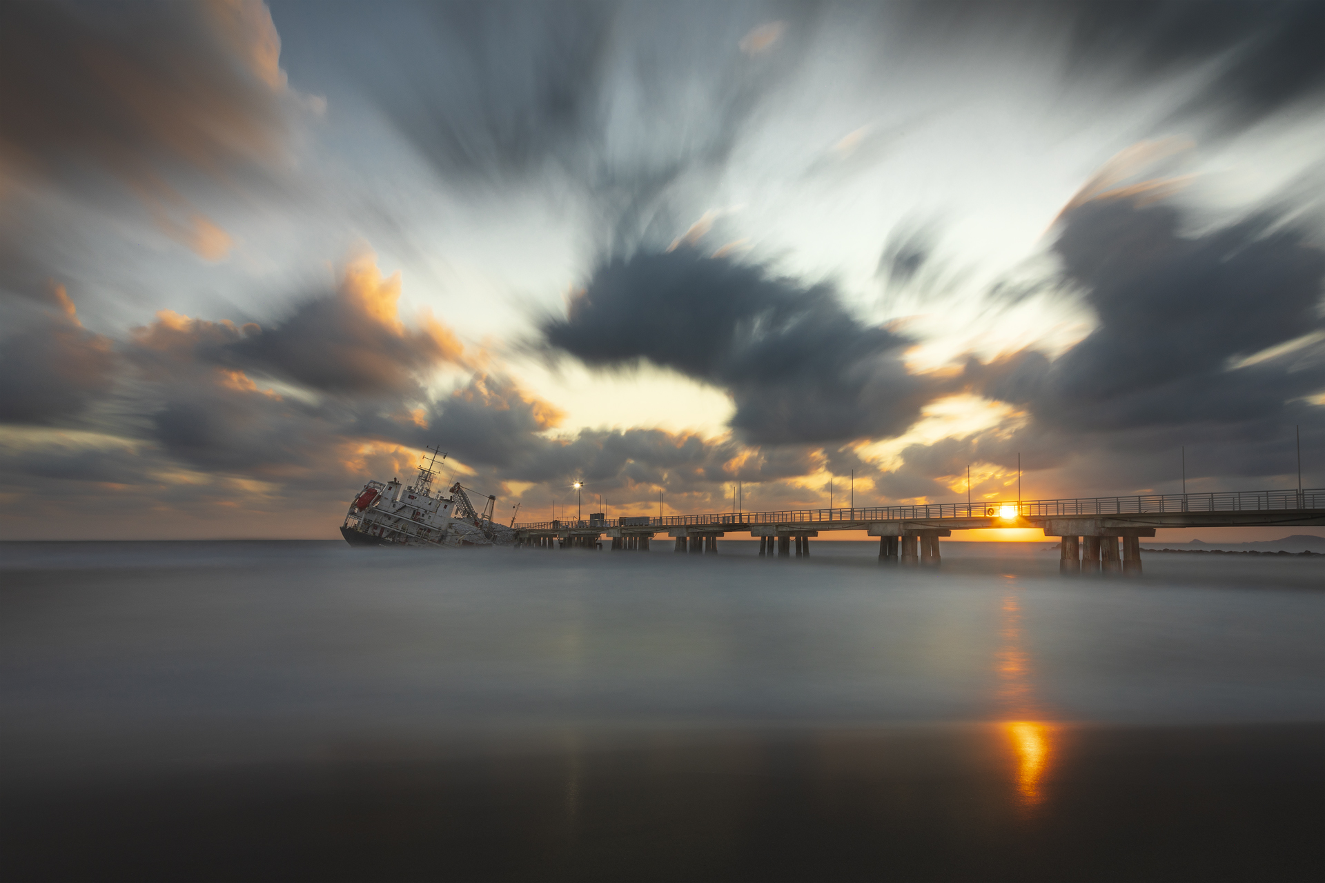 Fotografia di un relitto inclinato accanto a un pontile, con il sole che tramonta all’orizzonte e il cielo carico di nuvole.
